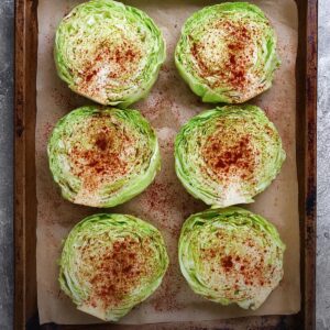 Easy Homemad Roasted Cabbage Steaks Recipe 16 Six raw cabbage steaks arranged neatly on a parchment-lined baking tray, each slice sprinkled with paprika and spices, overhead view, bright natural lighting, clean food-prep setting.