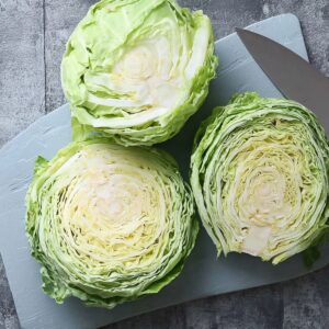 Easy Homemad Roasted Cabbage Steaks Recipe 15 Fresh cabbage cut into thick round slices on a light blue cutting board with a chef’s knife beside them. Top-down view, detailed texture of cabbage layers, neutral stone background.