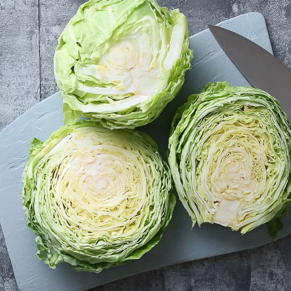 Easy Homemad Roasted Cabbage Steaks Recipe 10 Fresh cabbage cut into thick round slices on a light blue cutting board with a chef’s knife beside them. Top-down view, detailed texture of cabbage layers, neutral stone background.