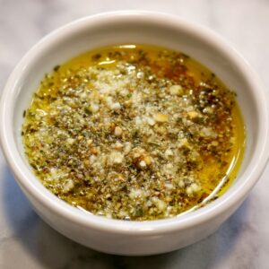 A close-up of a small bowl filled with melted butter mixed with minced garlic and Italian herbs, the oil forming a golden surface while the garlic pieces are visible.
