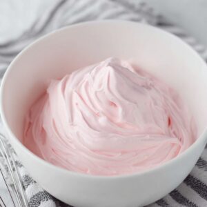 The Best Whipped Pink Lemonade Recipe 15 A close-up of fluffy pink whipped cream in a bowl, with a whisk placed next to the bowl on a striped cloth, suggesting the preparation process for the whipped topping.