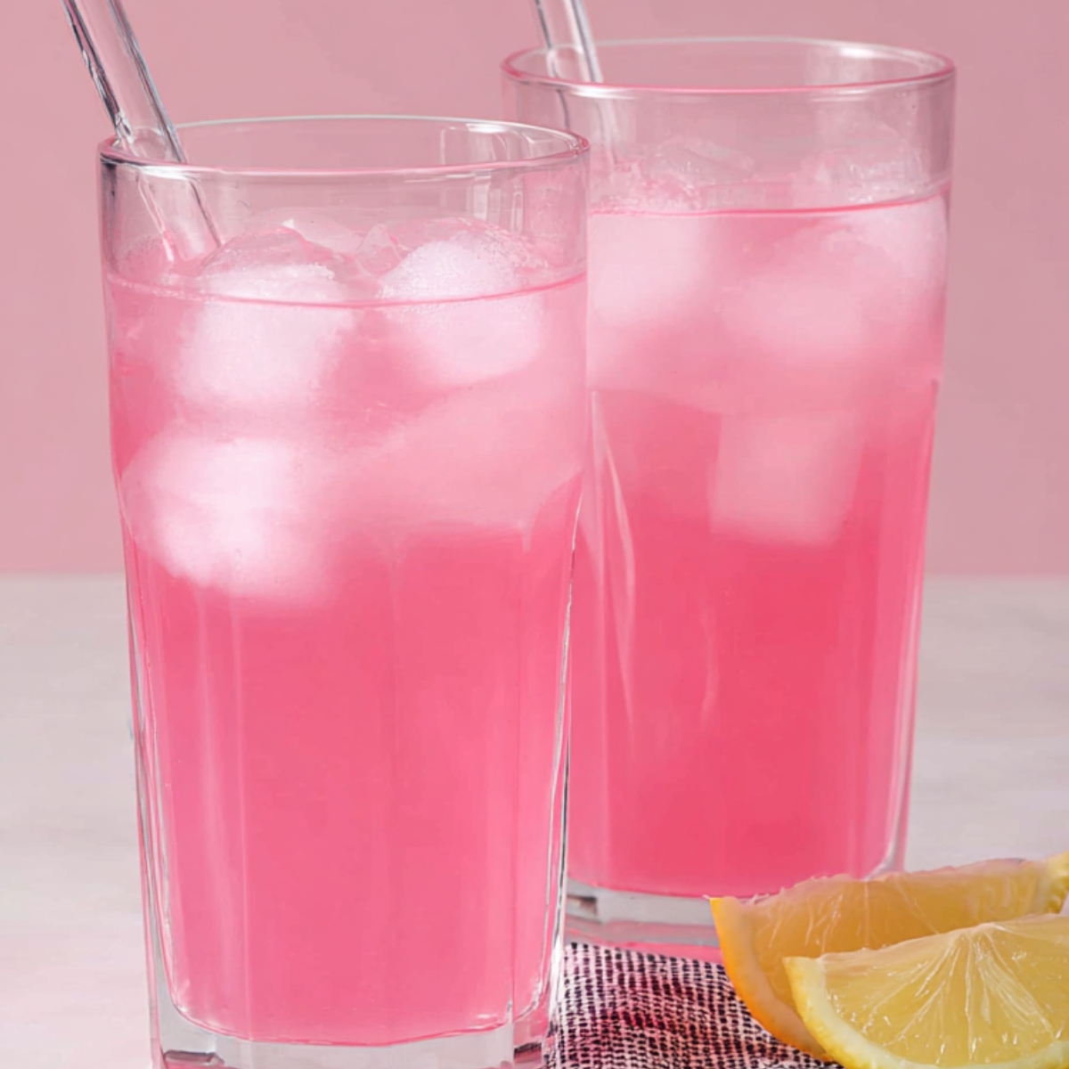 The Best Whipped Pink Lemonade Recipe 12 Two tall glasses of pink lemonade with ice, featuring clear straws. A few lemon slices are placed on the table beside the glasses. The background is a soft pink color.