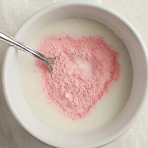 The Best Whipped Pink Lemonade Recipe 14 A close-up shot of a bowl containing pink powder, possibly a drink mix, resting on a soft cloth. A spoon is placed inside the bowl.