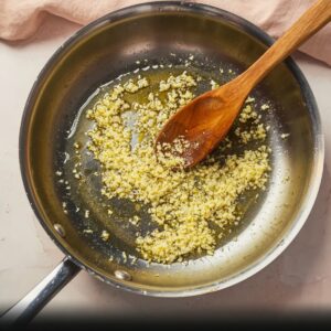 Best Gochujang Pasta Recipe | Creamy & Spicy in 20 Minutes 16 Minced garlic sautéing in olive oil in a stainless steel pan with a wooden spoon, close-up stovetop cooking scene.