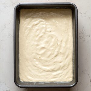 Easy Mango Tres Leches Recipe | Tropical Summer Dessert 17 Tres leches cake soaking in a creamy milk mixture inside a rectangular baking pan, photographed from above.