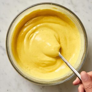 Easy Mango Tres Leches Recipe | Tropical Summer Dessert 18 Glass bowl filled with smooth mango lassi mixture being stirred with a spoon on a marble countertop.