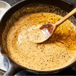 Easy Peppercorn Sauce Recipe | Ready in 20 Minutes 19 A wooden spoon stirring the creamy peppercorn sauce in a skillet, showing a smooth, velvety texture with black pepper bits throughout.