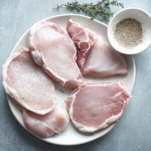 Easy Pineapple Pork Chops Recipe | Ready in 20 Minutes 15 Raw pork chops arranged on a white plate, lightly seasoned with salt and pepper, with herbs and spices nearby for preparation.