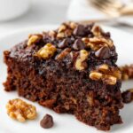 Chocolate Date Cake Close-up of a slice of chocolate date cake topped with chocolate chips and walnuts, served on a white plate, moist crumb, clean background, professional food photography.