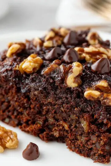 Best Chocolate Date Cake Recipe - Even Better Next Day 21 Chocolate Date Cake Close-up of a slice of chocolate date cake topped with chocolate chips and walnuts, served on a white plate, moist crumb, clean background, professional food photography.