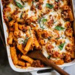 Baked Ziti Close-up of baked ziti pasta with melted mozzarella cheese, tomato meat sauce, and fresh basil, served from a white baking dish with a wooden spoon