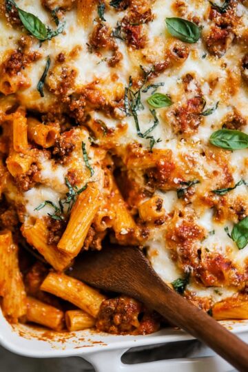 Best Baked Ziti Recipe | Feed a Crowd in Under 1 Hour 5 Baked Ziti Close-up of baked ziti pasta with melted mozzarella cheese, tomato meat sauce, and fresh basil, served from a white baking dish with a wooden spoon