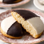 Black and White Cookies sliced open on a patterned plate, half covered in chocolate icing and half in vanilla icing.