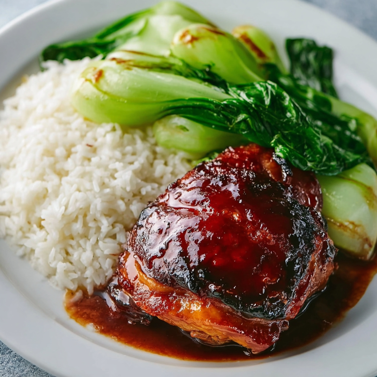 Char Siu Chicken Glazed char siu chicken thigh served with steamed white rice and sautéed bok choy on a white plate, coated in sticky red barbecue sauce.