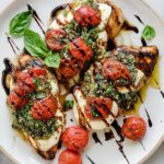 Chicken Caprese Overhead view of plated caprese chicken topped with melted mozzarella, roasted cherry tomatoes, basil pesto, and balsamic glaze on a white plate.