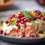 Christmas Salmon Recipe Extreme close-up of flaky baked salmon texture topped with creamy sauce, pomegranate arils, pistachio pieces, and fresh dill, shallow depth of field, soft bokeh background, luxurious food photography, vibrant colors, high detail.”