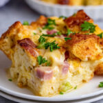 Eggs Benedict Casserole Close-up of a baked Eggs Benedict casserole slice with golden bread cubes, creamy egg filling, ham, and parsley garnish on a white plate.