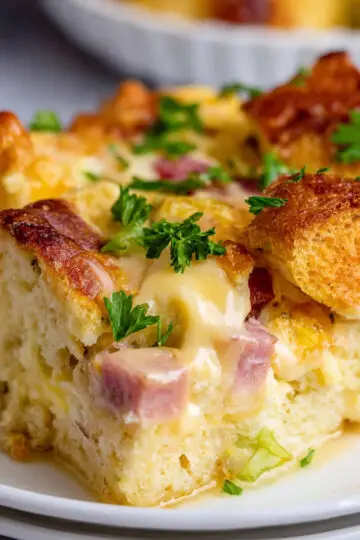 Overnight Eggs Benedict Casserole Recipe | Feeds 12 1 Eggs Benedict Casserole Close-up of a baked Eggs Benedict casserole slice with golden bread cubes, creamy egg filling, ham, and parsley garnish on a white plate.