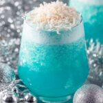 Jack Frost Cocktail A close-up shot of a festive blue cocktail topped with shredded coconut, surrounded by silver holiday decorations such as ornaments and tinsel."