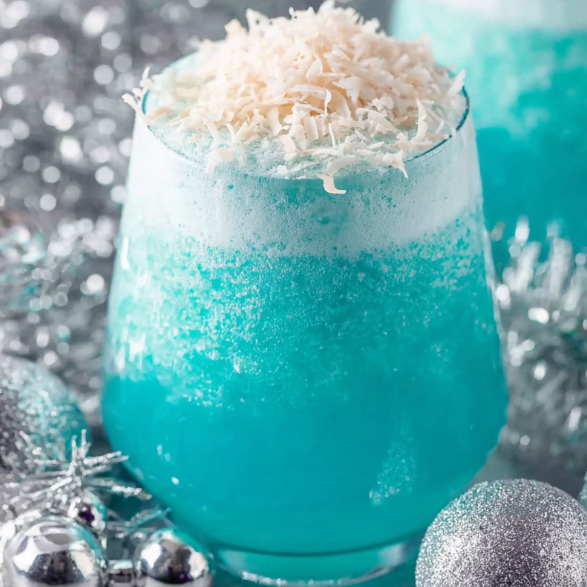 Jack Frost Cocktail A close-up shot of a festive blue cocktail topped with shredded coconut, surrounded by silver holiday decorations such as ornaments and tinsel."