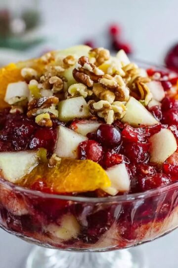 Jello Fruit Salad Recipe | Easy 10-Ingredient Classic 2 Jello Fruit Salad A beautifully presented fruit salad with cranberries, topped with orange slices, pear pieces, and chopped nuts in a clear glass bowl, perfect for a festive occasion.