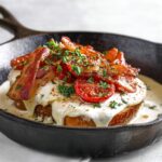 Kentucky Hot Brown Hot brown sandwich served in a cast iron skillet with thick toast, creamy sauce, crispy bacon strips, roasted tomatoes, and chopped herbs