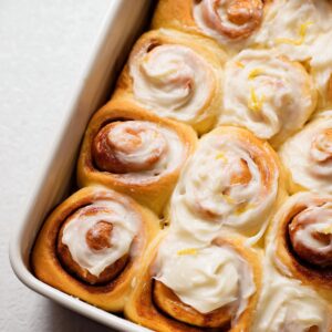 Lemon Cinnamon Rolls Recipe | Soft, Tangy & Easy 19 Overhead close-up of freshly baked lemon sweet rolls in a white baking dish, generously frosted with lemon cream cheese icing, golden dough swirls visible, bright airy lighting, clean bakery aesthetic, high-resolution food photography