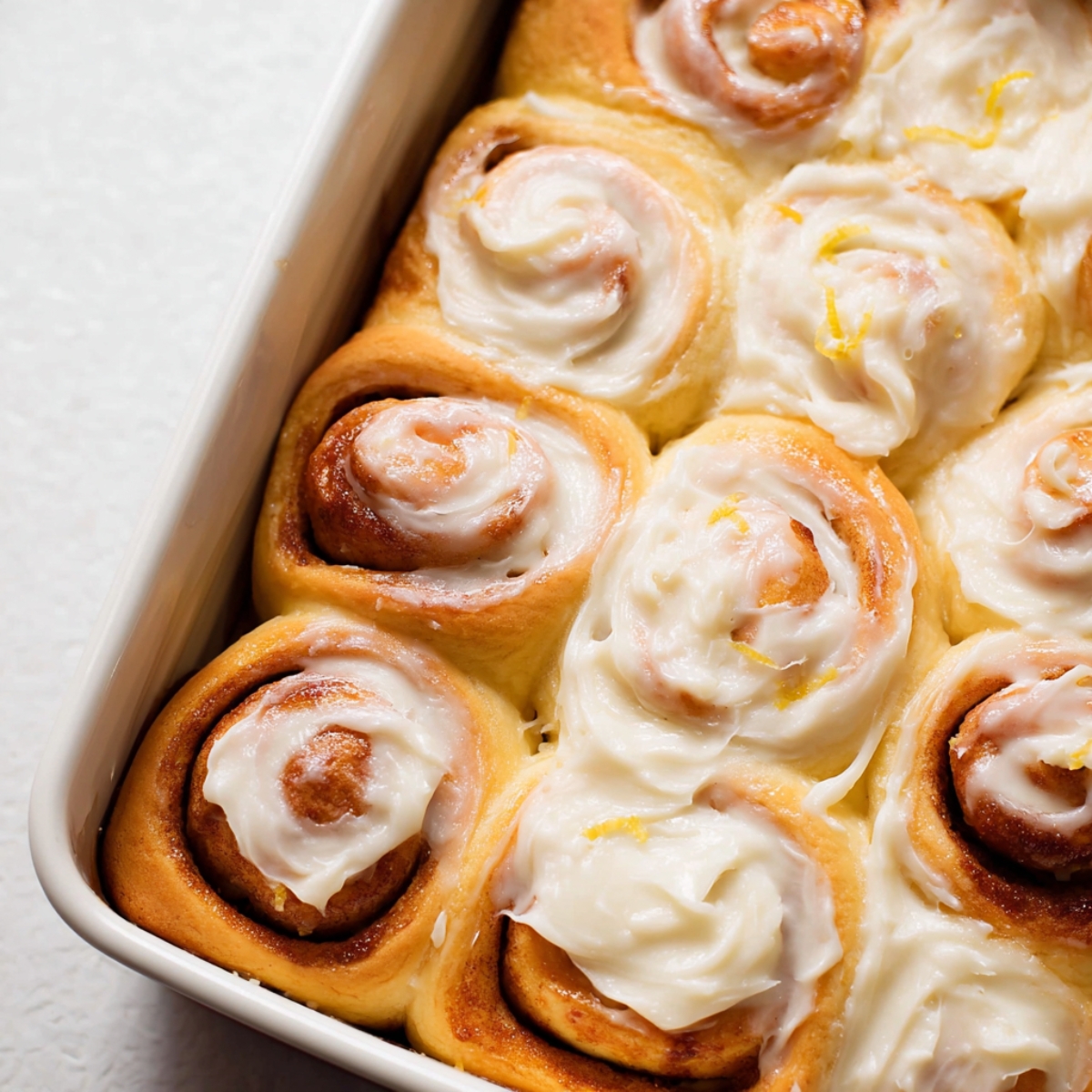 Lemon Cinnamon Rolls Recipe | Soft, Tangy & Easy 13 Overhead close-up of freshly baked lemon sweet rolls in a white baking dish, generously frosted with lemon cream cheese icing, golden dough swirls visible, bright airy lighting, clean bakery aesthetic, high-resolution food photography