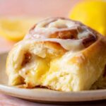 Lemon Cinnamon Rolls Close-up food photography of a soft lemon sweet roll cut open, filled with gooey lemon sugar filling and topped with glossy lemon cream cheese frosting, bright warm lighting, shallow depth of field, bakery-style dessert, ultra realistic, high detail, soft pastel background