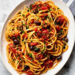 Pasta Puttanesca Overhead view of spaghetti alla puttanesca in a white oval dish, with long noodles coated in tomato sauce, black olives, capers, chopped parsley, and red pepper flakes on a light marble surface.