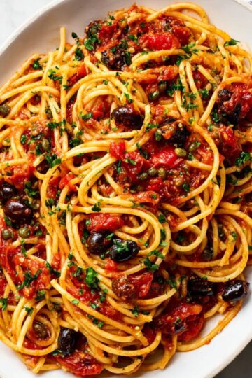 Pasta Puttanesca Recipe | Quick & Easy in 30 Minutes 7 Pasta Puttanesca Overhead view of spaghetti alla puttanesca in a white oval dish, with long noodles coated in tomato sauce, black olives, capers, chopped parsley, and red pepper flakes on a light marble surface.