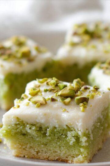 Pistachio Cookie Bars Close-up of pistachio cookie bars cut into squares on a white plate, soft green interior with creamy white frosting on top, sprinkled chopped pistachios, natural window light, shallow depth of field, elegant homemade dessert photography, cozy and minimal styling.