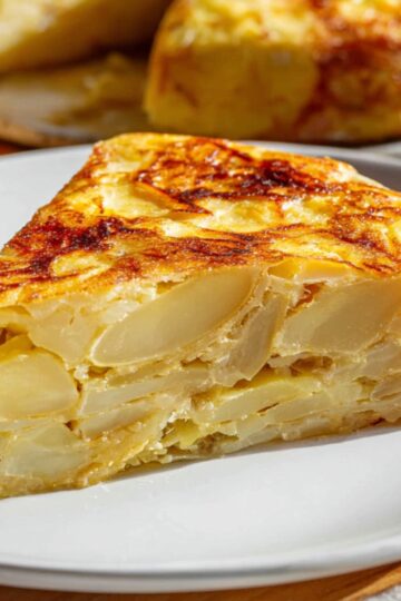 Spanish Tortilla Recipe - Easy 5-Ingredient Authentic 6 Spanish tortilla Close-up of a slice of Spanish tortilla on a white plate with bread, layered potatoes visible inside, golden crust, warm natural light, Mediterranean dish photography