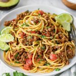 Plated taco spaghetti with seasoned ground beef, tomatoes, and herbs, served with lime wedges and garnished with cilantro.