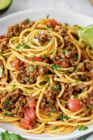 One-Pan Taco Spaghetti Recipe | Ready in 30 Minutes 4 Plated taco spaghetti with seasoned ground beef, tomatoes, and herbs, served with lime wedges and garnished with cilantro.