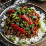 Thai Basil Beef A bowl of Thai basil beef served on rice, garnished with green onions and fresh basil leaves."
