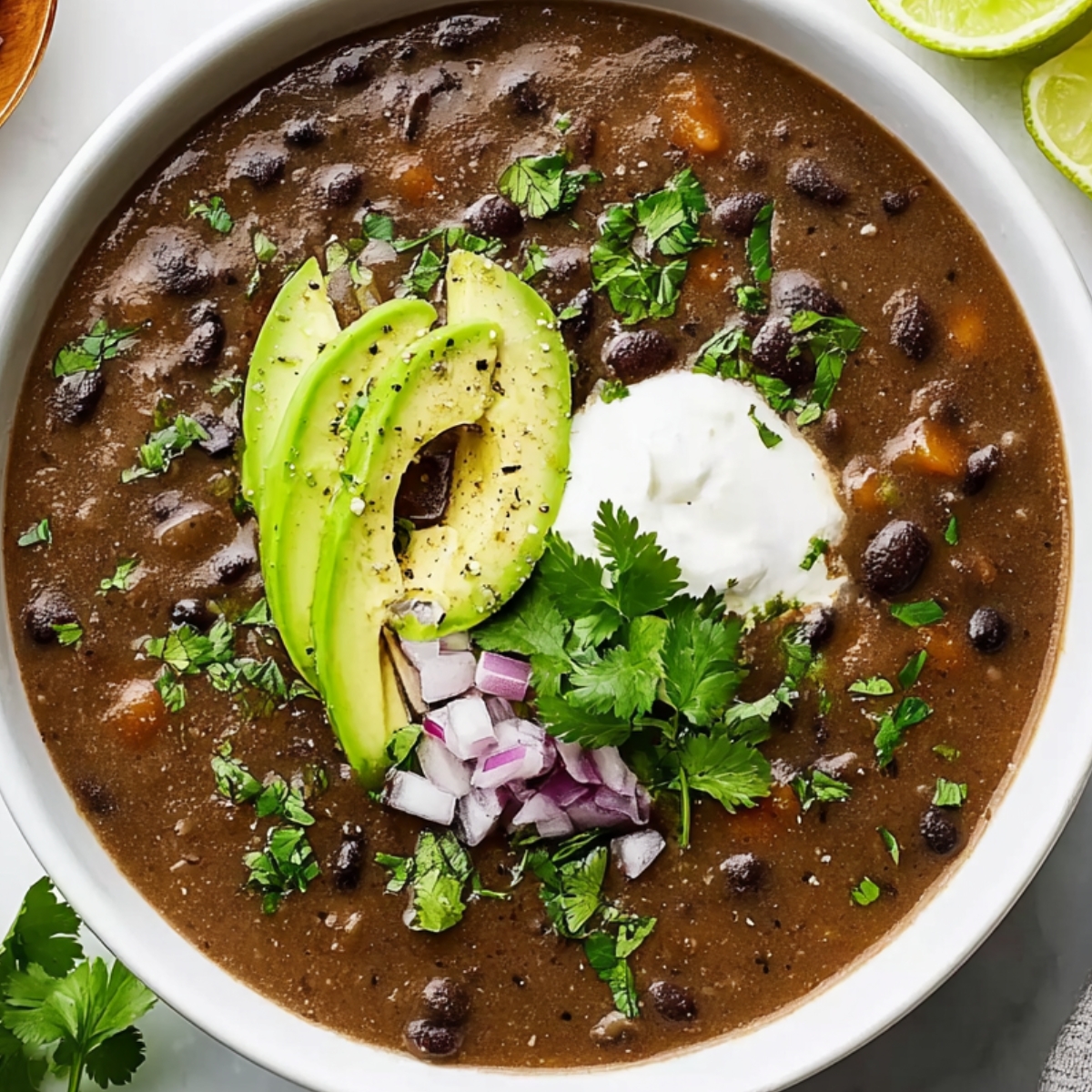 Top-down photo of creamy black bean soup in a white bowl, topped with avocado slices, sour cream, chopped red onion, and fresh cilantro, lime wedges on the side, bright natural light, clean white background