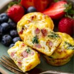 breakfast egg muffins A plate of egg muffins served with fresh strawberries and blueberries, showing a close-up of the muffins cut in half to reveal their filling of ham, spinach, and cheese.