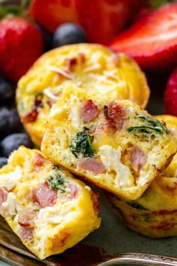 Healthy Breakfast Egg Muffins Recipe 16 breakfast egg muffins A plate of egg muffins served with fresh strawberries and blueberries, showing a close-up of the muffins cut in half to reveal their filling of ham, spinach, and cheese.