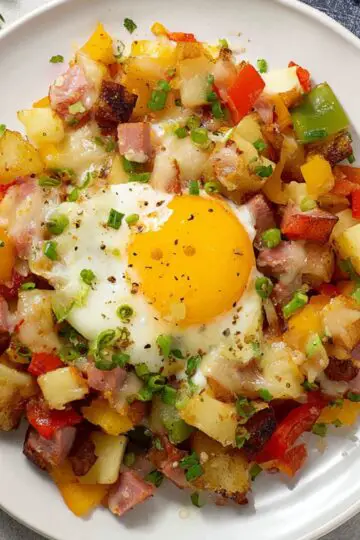 30-Minute Breakfast Skillet | Crispy Potatoes & Eggs 8 breakfast skillet hash on a white plate with crispy potatoes, diced ham, red and green bell peppers, melted cheese, and a sunny-side-up egg on top.