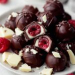 chocolate covered raspberries on a white plate, some cut open to reveal fresh red raspberry centers, garnished with white chocolate pieces.