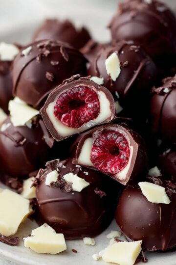 The Best Chocolate Covered Raspberries Recipe 12 chocolate covered raspberries on a white plate, some cut open to reveal fresh red raspberry centers, garnished with white chocolate pieces.