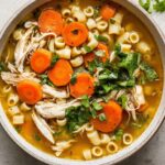 ginger chicken soup Overhead view of a bowl of homemade chicken noodle soup with shredded chicken, sliced carrots, small pasta, and fresh herbs in a golden broth.
