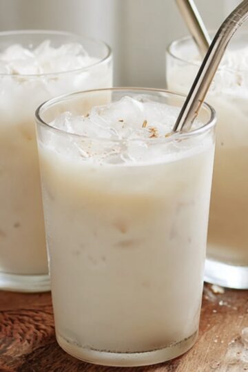 Authentic Horchata Recipe | Homemade Mexican Drink 6 horchata recipe Three small clear glasses filled with creamy Mexican horchata over ice, placed on a rustic carved wooden board, stainless steel reusable straws in the glasses, soft natural window light, white minimal background, elegant food photography, airy and bright mood, shallow depth of field, high detail, lifestyle drink photography, neutral tones, 50mm lens, f/2.8