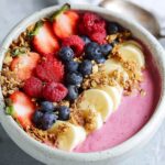 raspberry smoothie bowl Raspberry smoothie bowl topped with granola, fresh berries, and banana slices, placed on a marble countertop.