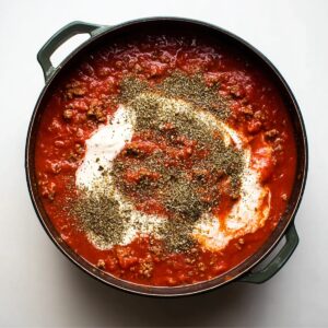 Best Baked Ziti Recipe | Feed a Crowd in Under 1 Hour 15 Overhead view of tomato meat sauce topped with cream and dried Italian herbs in a dark pot on a white background