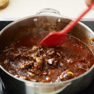Easy Beef Mechado Recipe - Tender Filipino Tomato Beef Stew 17 Thick beef mechado sauce being stirred in a stainless steel pot with a red spatula during cooking.