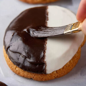 Black and White Cookies | Easy Homemade Recipe Guide 17 Close-up of a black and white cookie as chocolate icing is being spread on one half with a small spatula.