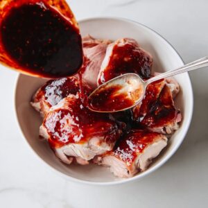 Easy Char Siu Chicken Recipe | Sticky Chinese BBQ Chicken 16 Raw chicken thighs in a bowl being coated with thick char siu marinade, with dark red sauce poured and spread using a spoon.