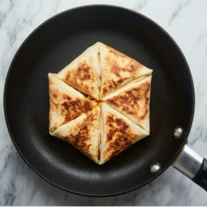 Chicken Crunch Wrap Recipe - Crispy & Ready in 30 Min 16 Top-down food photography of a folded tortilla wrap being pan-fried in a black nonstick skillet, golden brown crispy surface with triangular folds, minimal styling, marble countertop background, natural soft lighting, clean cooking process, high realism
